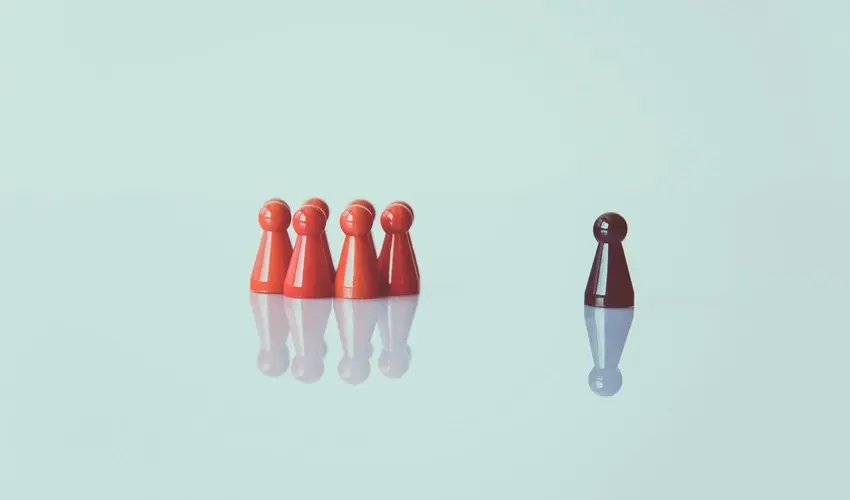 Five red game pieces grouped together facing a single darker piece standing apart on a reflective surface.